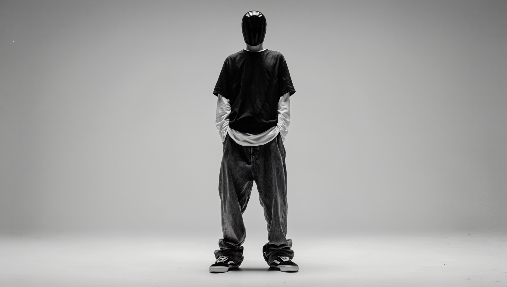 Ein Person steht vor einem schlichten grauen Hintergrund. Sie trägt ein schwarzes T-Shirt über einem langärmeligen weißen Shirt, weite schwarze Hosen und schwarze Sneaker. Ihr Gesicht ist mit einer glänzenden schwarzen Maske verdeckt. Die Pose wirkt entspannt, mit den Händen in den Hosentaschen.