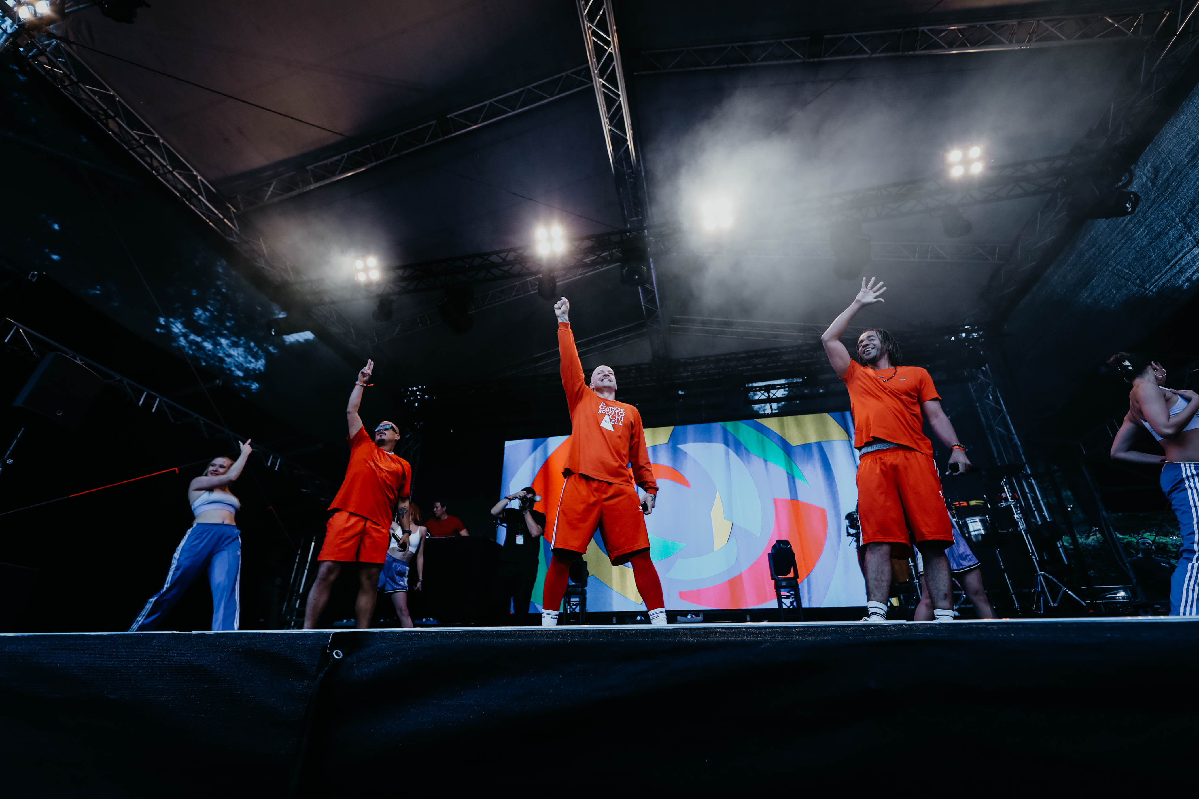 Culcha Candela Bandmitglieder auf Bühne, tragen orangefarbene Outfits, heben Arme während Auftritts.