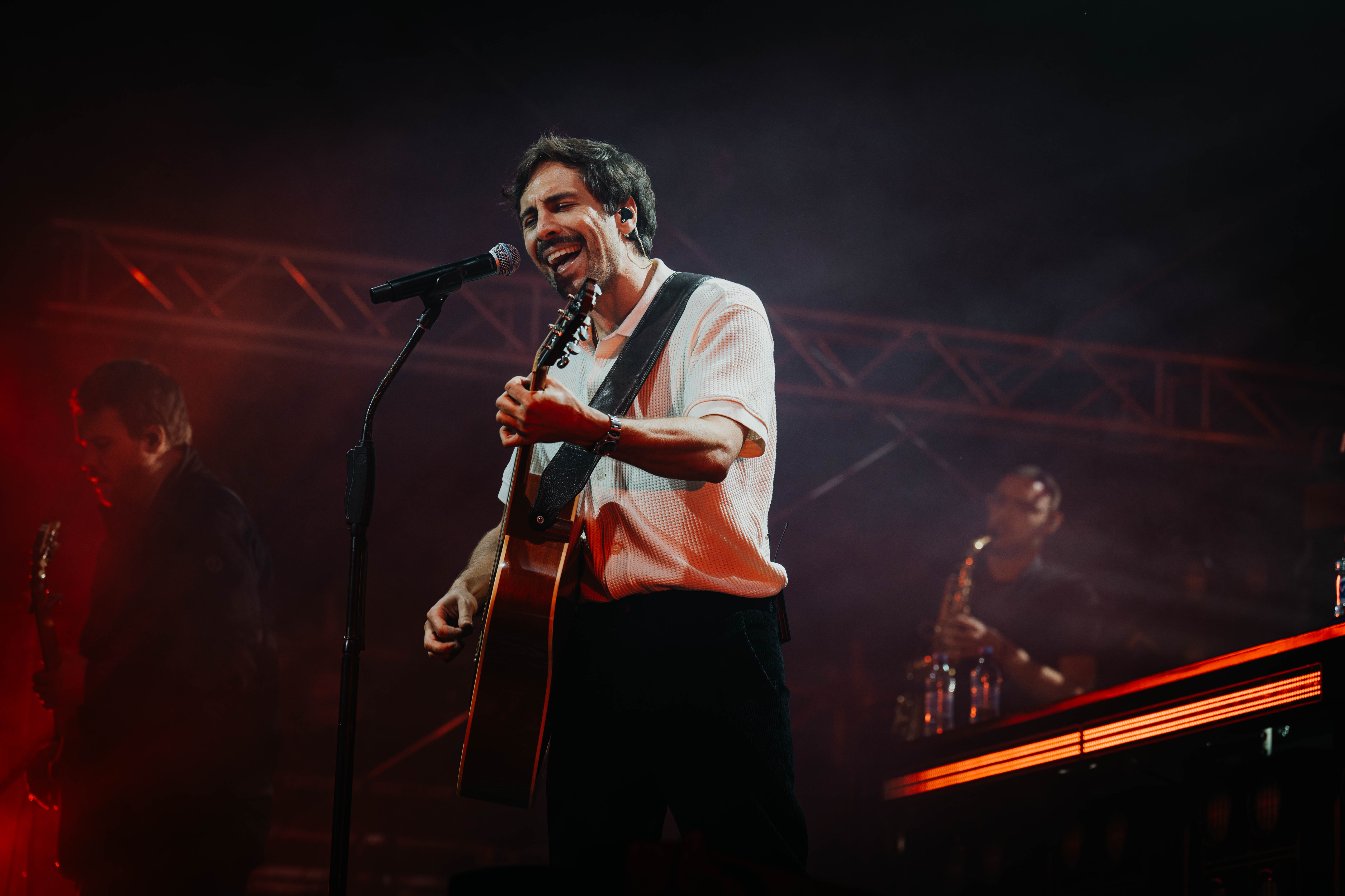 Max Giesinger spielt Gitarre und singt am Mikrofon während eines Konzerts mit roter Bühnenbeleuchtung.
