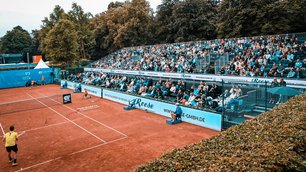 Ein Tennisplatz mit rotem Sandbelag, auf dem zwei Spieler ein Match bestreiten. Am Spielfeldrand sitzt ein Schiedsrichter. Die Tribünen sind gut gefüllt mit Zuschauern, die das Spiel verfolgen. Im Hintergrund sind dicht bewachsene Bäume zu sehen. Schilder mit Werbung sind entlang des Zauns und auf den Tribünen gut sichtbar angebracht.