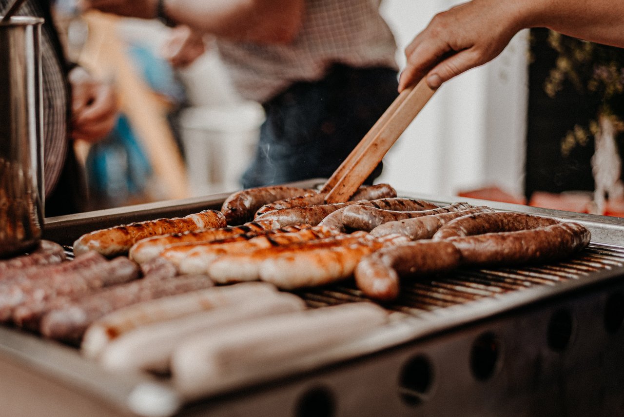 Eine Person grillt verschiedene Sorten von Würstchen auf einem Grill. Einige Würstchen sind bereits gebräunt, während andere noch heller sind. Die Person wendet eine Wurst mit einer Grillzange oder einem Holzspatel. Im Hintergrund sind unscharf weitere Personen zu erkennen. Der Grill befindet sich vermutlich im Freien.