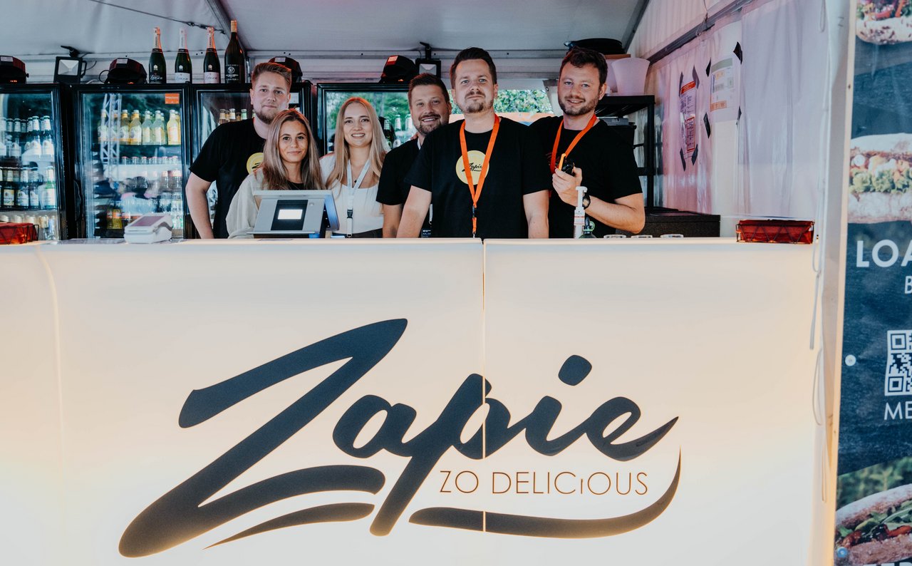 Fünf Personen hinter einem Verkaufsstand mit dem Schriftzug "Zapie Zo Delicious", tragen schwarze und weiße Kleidung, einige mit orangen Lanyards.