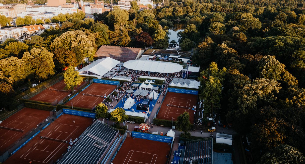 Luftaufnahme eines Tennisplatzes mit Zuschauern und Zelten, umgeben von Bäumen und Gebäuden im Hintergrund.