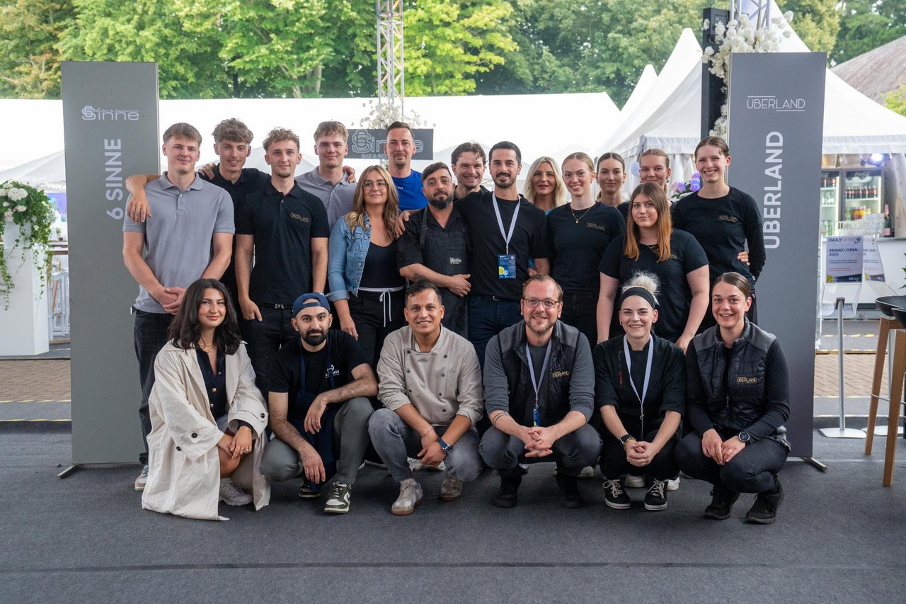 Auf dem Bild ist eine Gruppe von 19 Personen, die zusammen posiert. Sie stehen und knien in zwei Reihen vor einem Hintergrund mit weißen Zelten und Bäumen. Die Gruppe wirkt freundlich und ist überwiegend in Freizeit- und Arbeitskleidung gekleidet. Seitlich sind zwei Schilder mit den Aufschriften "6 Sinne" und "Überland" sichtbar.