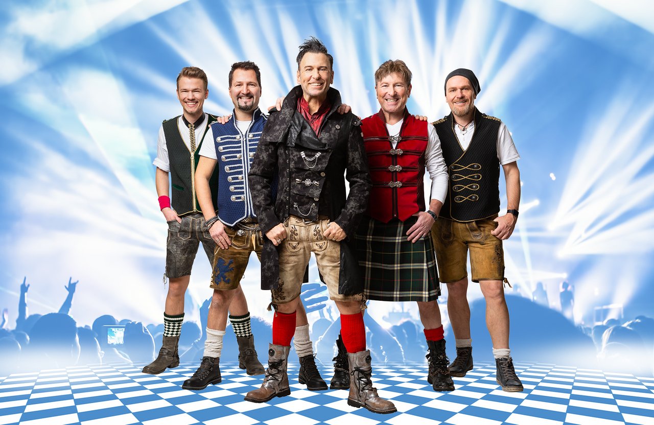 Fünf Männer stehen auf einem blau-weiß karierten Boden vor einer beleuchteten Bühne. Sie tragen moderne Trachtenmode, darunter Lederhosen, Westen und Stiefel. Der Hintergrund zeigt Lichteffekte und die Silhouetten eines Publikums. Alle lächeln in die Kamera.