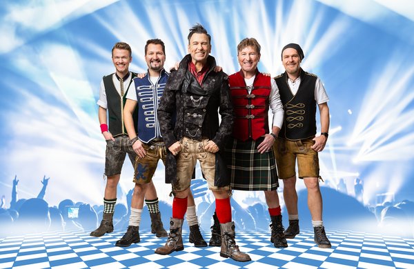 Fünf Männer stehen auf einem blau-weiß karierten Boden vor einer beleuchteten Bühne. Sie tragen moderne Trachtenmode, darunter Lederhosen, Westen und Stiefel. Der Hintergrund zeigt Lichteffekte und die Silhouetten eines Publikums. Alle lächeln in die Kamera.
