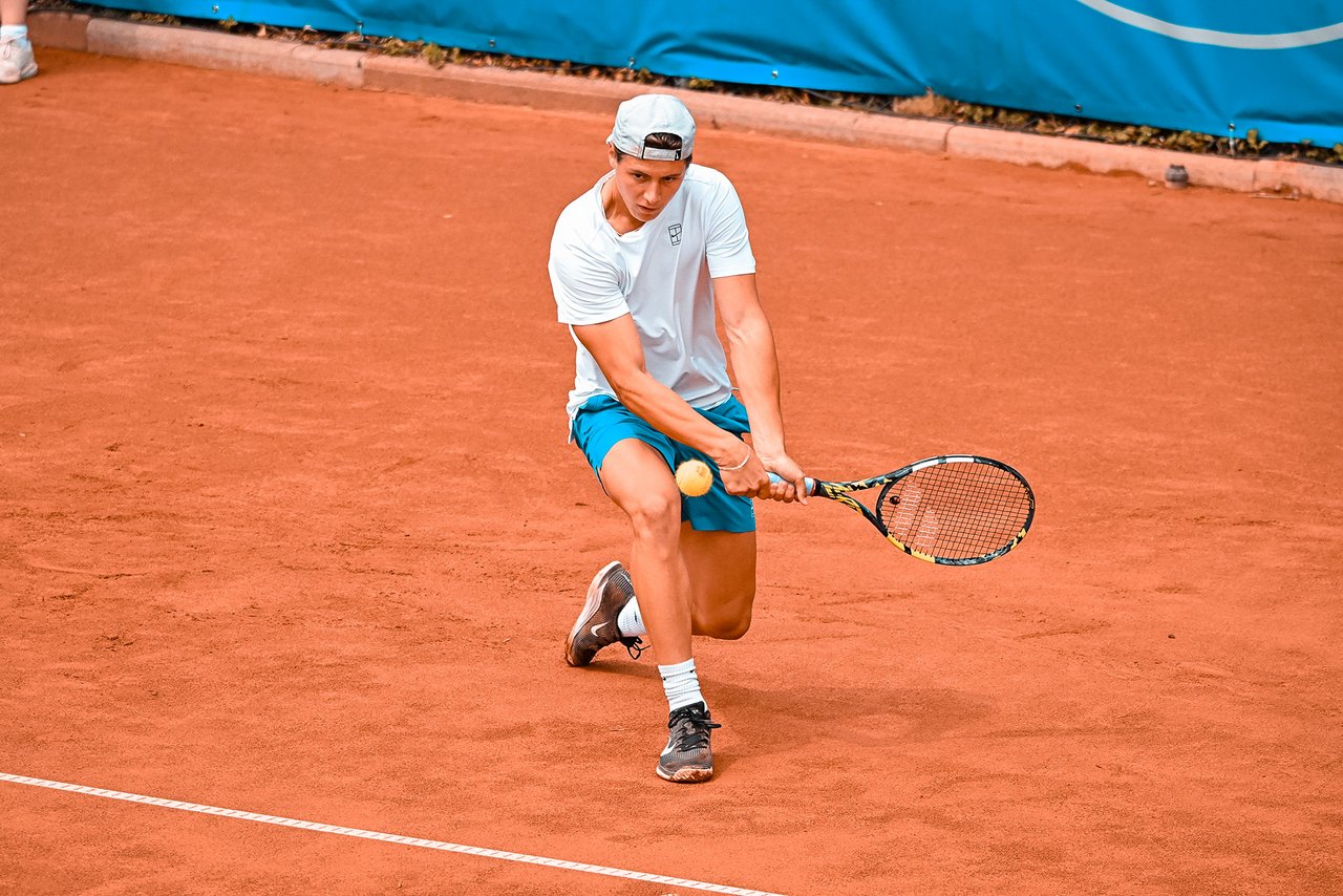 Tennisspieler in weißem T-Shirt, blauer Shorts und weißer Kappe spielt Rückhand auf Sandplatz.