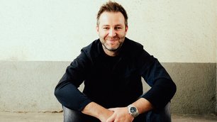Ein Mann sitzt auf einem Bordstein und lächelt in die Kamera. Er trägt einen schwarzen Pullover, Jeans und braune Stiefel. Im Hintergrund ist eine beige Wand zu sehen. Auf dem Boden liegt ein Papierstück.