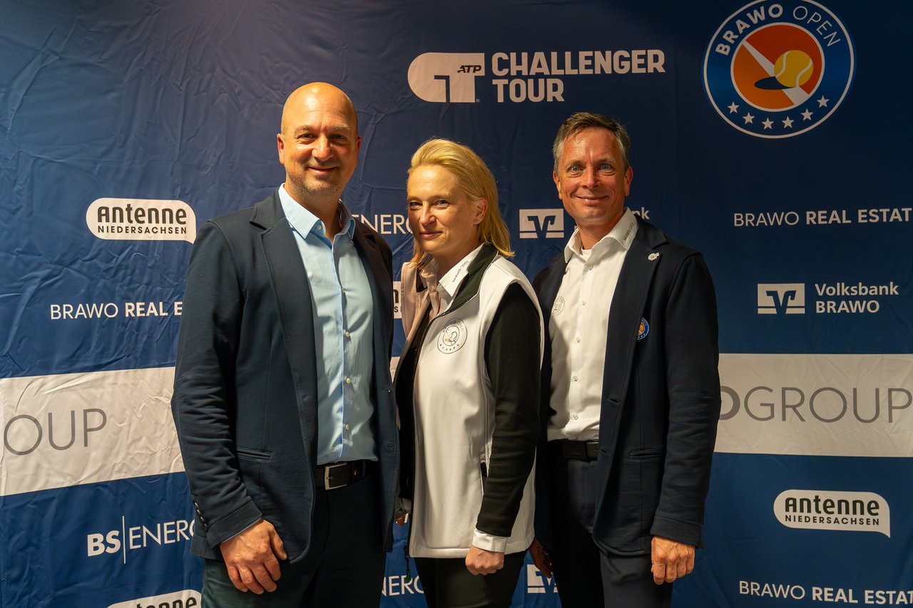 Drei Personen stehen vor einem blauen Hintergrund mit verschiedenen Sponsorenlogos, darunter "ATP Challenger Tour" und "Brawo Open". Die Person links trägt einen dunklen Anzug mit hellblauem Hemd. In der Mitte befindet sich eine Person mit blonder Frisur, die eine weiße Weste über einem Hemd trägt. Rechts steht eine weitere Person in einem dunklen Anzug mit weißem Hemd. Alle drei lächeln in die Kamera.