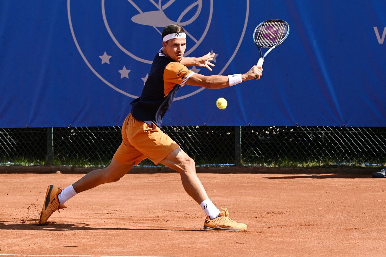 Tennisspieler in orangefarbener Sportkleidung schlägt auf Sandplatz mit ausgestrecktem Arm einen Ball mit dem Schläger zurück.