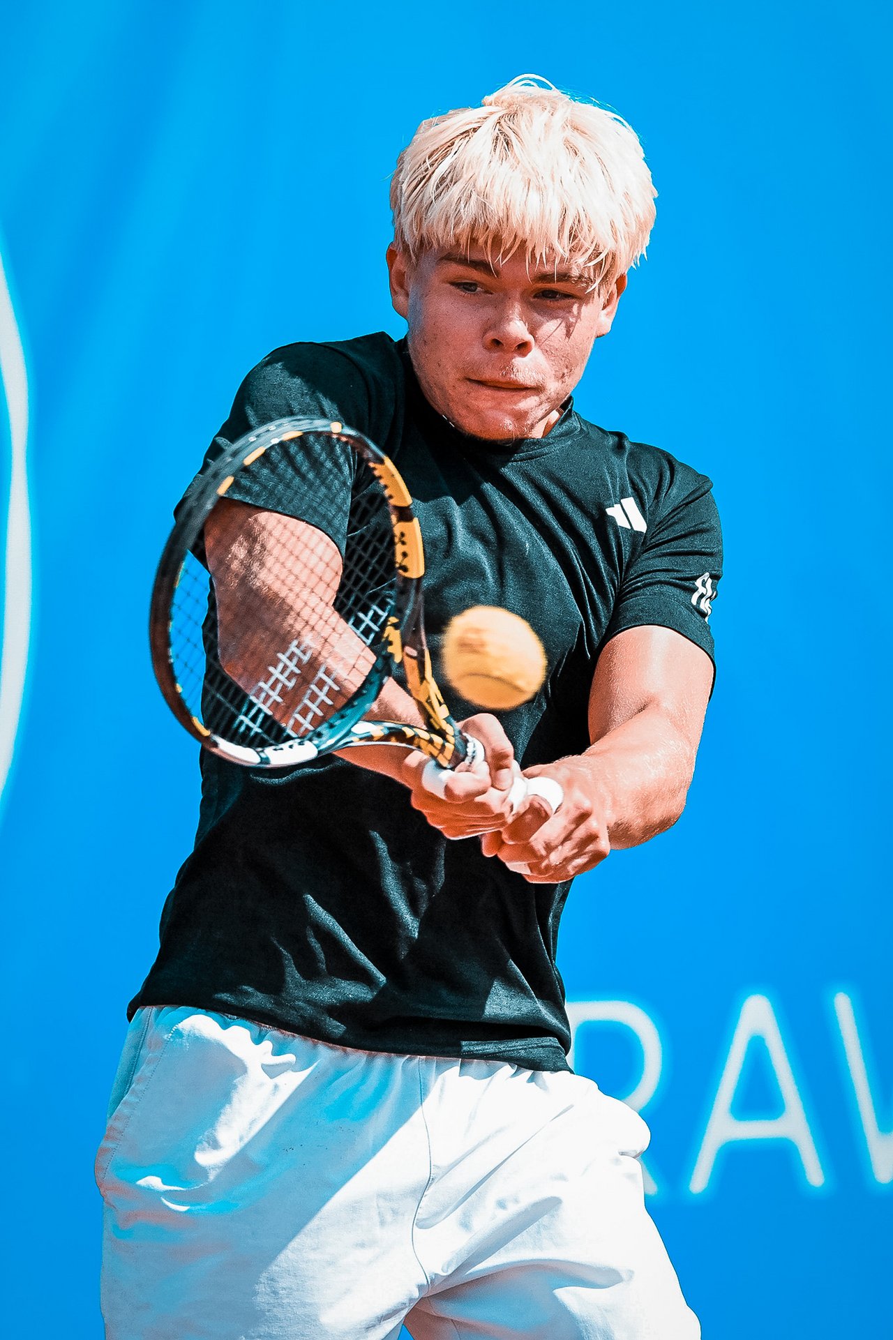 Tennisspieler mit blondem Haar in schwarzem T-Shirt und weißer Hose schlägt Ball mit Tennisschläger vor blauem Hintergrund.