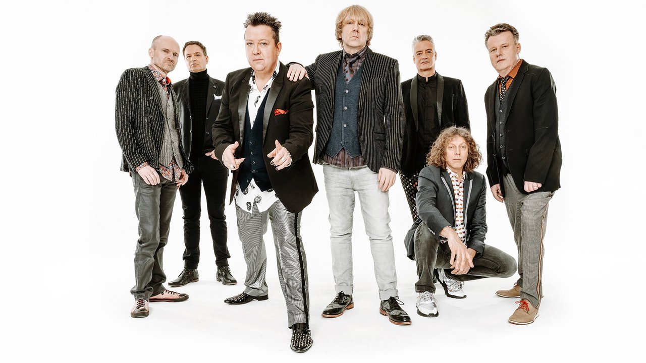 Die Prinzen Eine Gruppe von sieben Männern posiert in einem Fotostudio vor einem weißen Hintergrund. Sie tragen verschiedene stilvolle Anzüge und Hemden, die kreativ und individuell gestaltet sind. Einige stehen, während einer in der Hocke sitzt. Die Gesichtsausdrücke sind selbstbewusst, und die meisten schauen direkt in die Kamera.