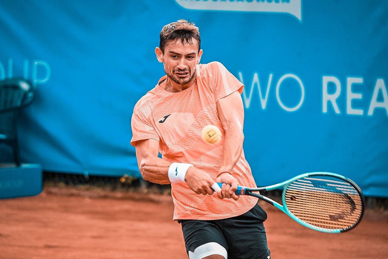 Tennisspieler Mariano Navone in orangefarbenem Shirt schlägt Rückhand auf Sandplatz, Ball vor Schläger.