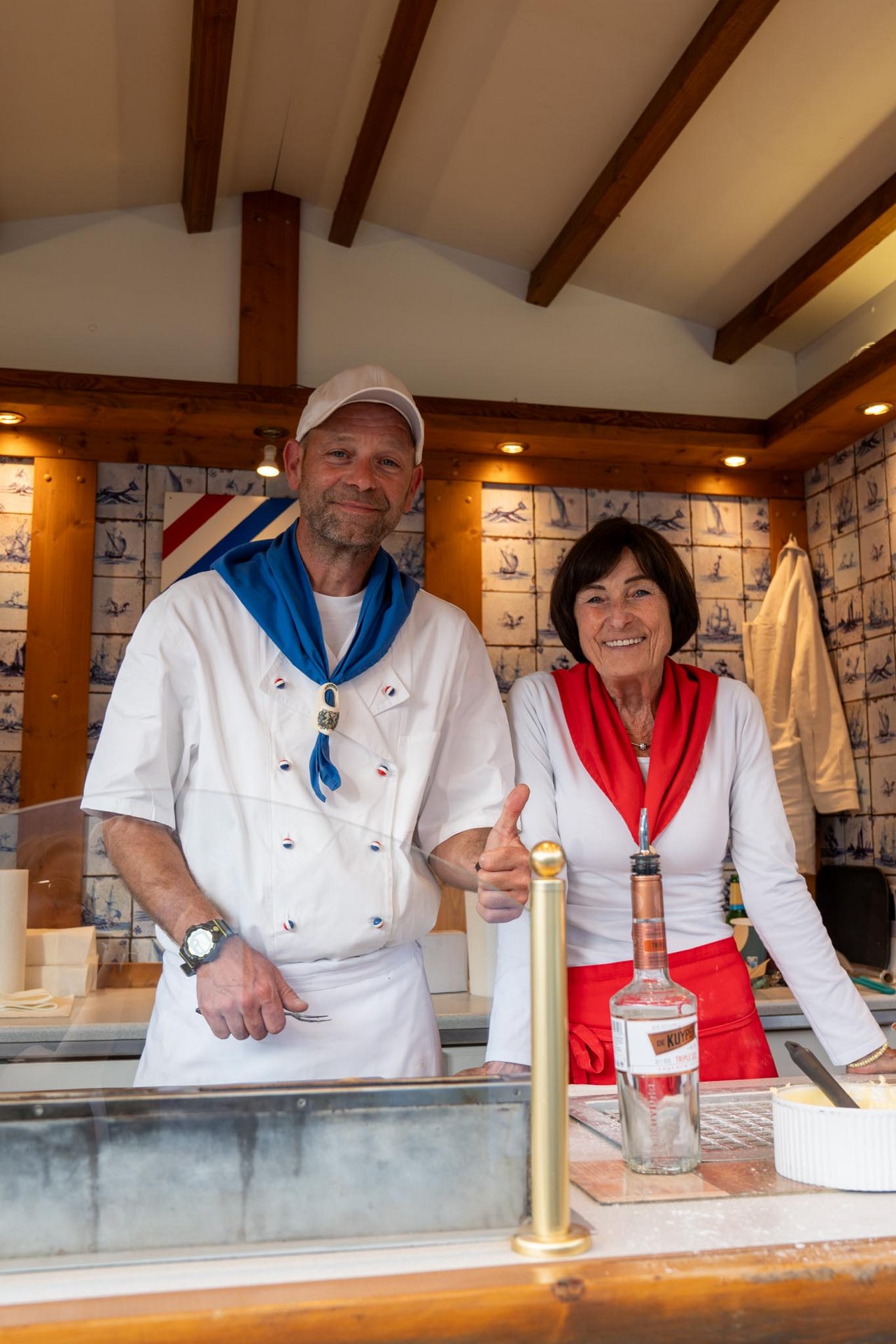 Ein Mann und eine Frau stehen in einem Verkaufsstand und lächeln in die Kamera. Der Mann trägt eine weiße Kochuniform, eine blaue Halstuch und eine weiße Mütze. Die Frau trägt ein weißes Oberteil und ein rotes Halstuch. Vor ihnen befindet sich eine Theke mit einer Flasche und weiteren Gegenständen. Die Wände des Stands sind mit Kacheln dekoriert.