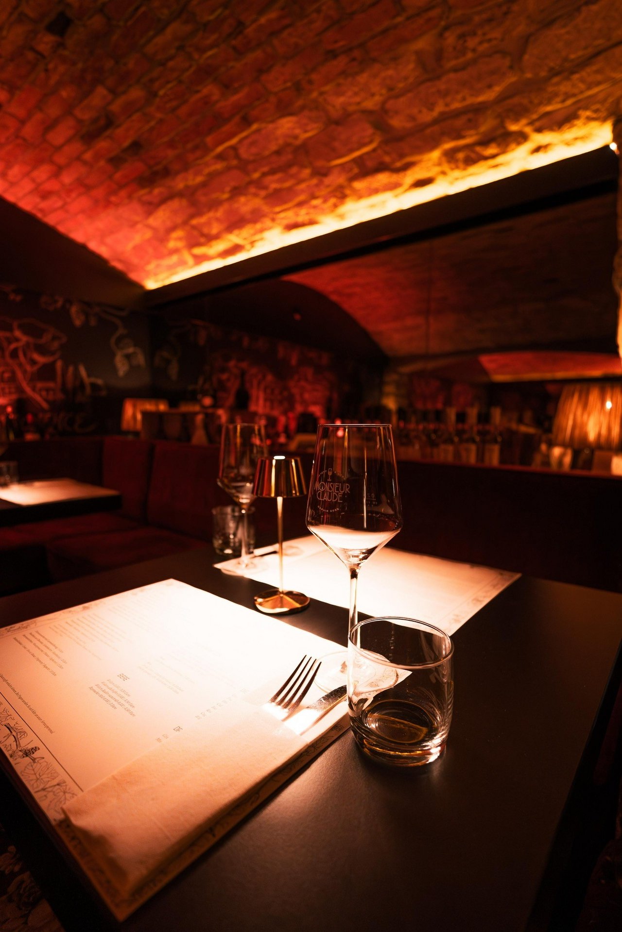 Ein stilvoll beleuchtetes Restaurant mit Gewölbedecke und warmer, rötlicher Beleuchtung. Auf einem dunklen Tisch stehen eingedeckt ein Weinglas mit Gravur und ein leeres Wasserglas. Neben dem Glas liegt ein Menü unter einem Stoffserviette, das teilweise sichtbar ist. Im Hintergrund befinden sich weitere Tische und eine künstlerisch gestaltete Wand.