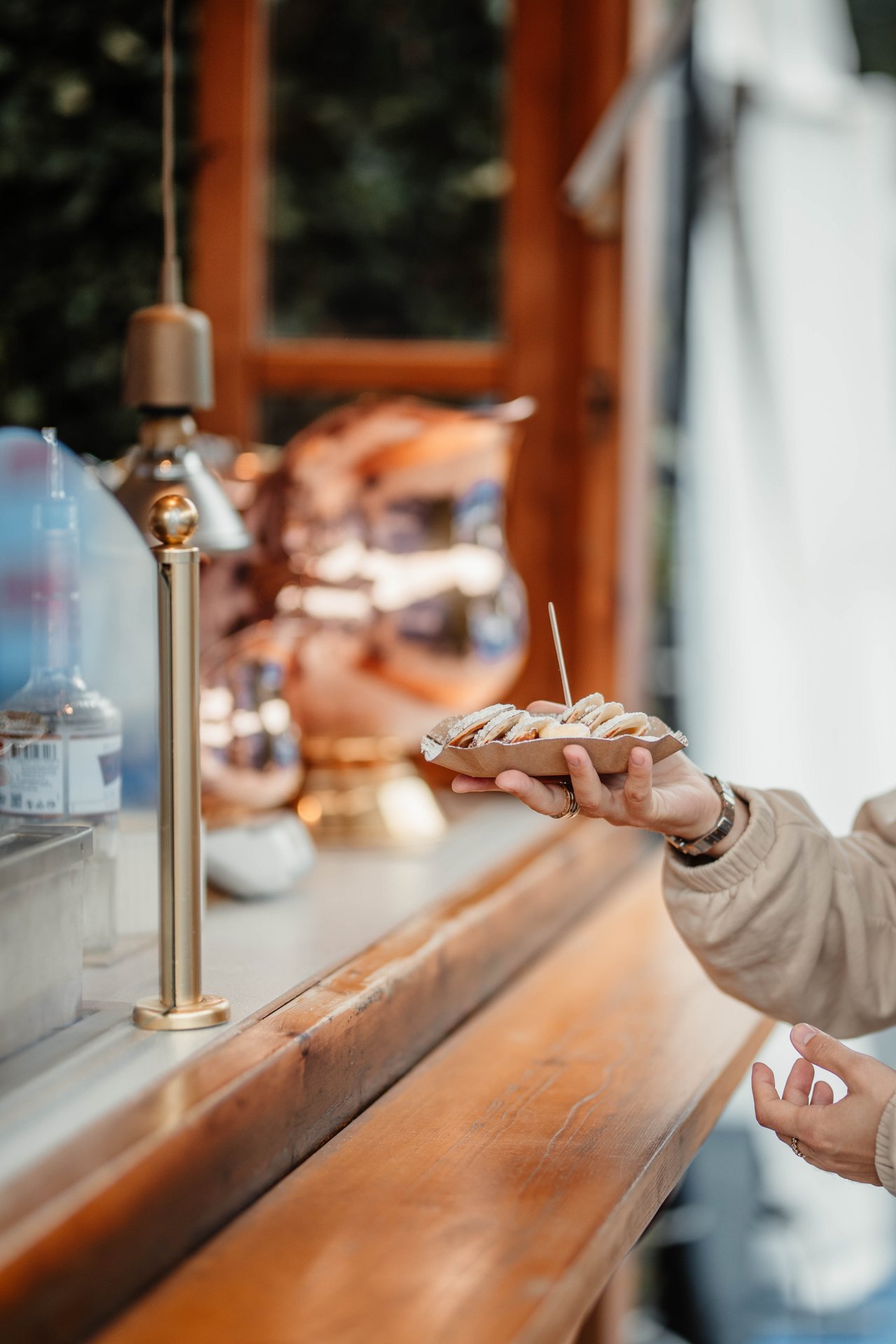 Ein Foto zeigt eine Hand, die eine kleine Pappschale mit frischen Waffeln hält. Im Hintergrund sieht man einen hölzernen Stand und glänzendes Kupfergeschirr. Eine Person trägt eine beige Jacke und hat Schmuck an den Fingern. Ein Zahnstocher ragt aus den Waffeln heraus.