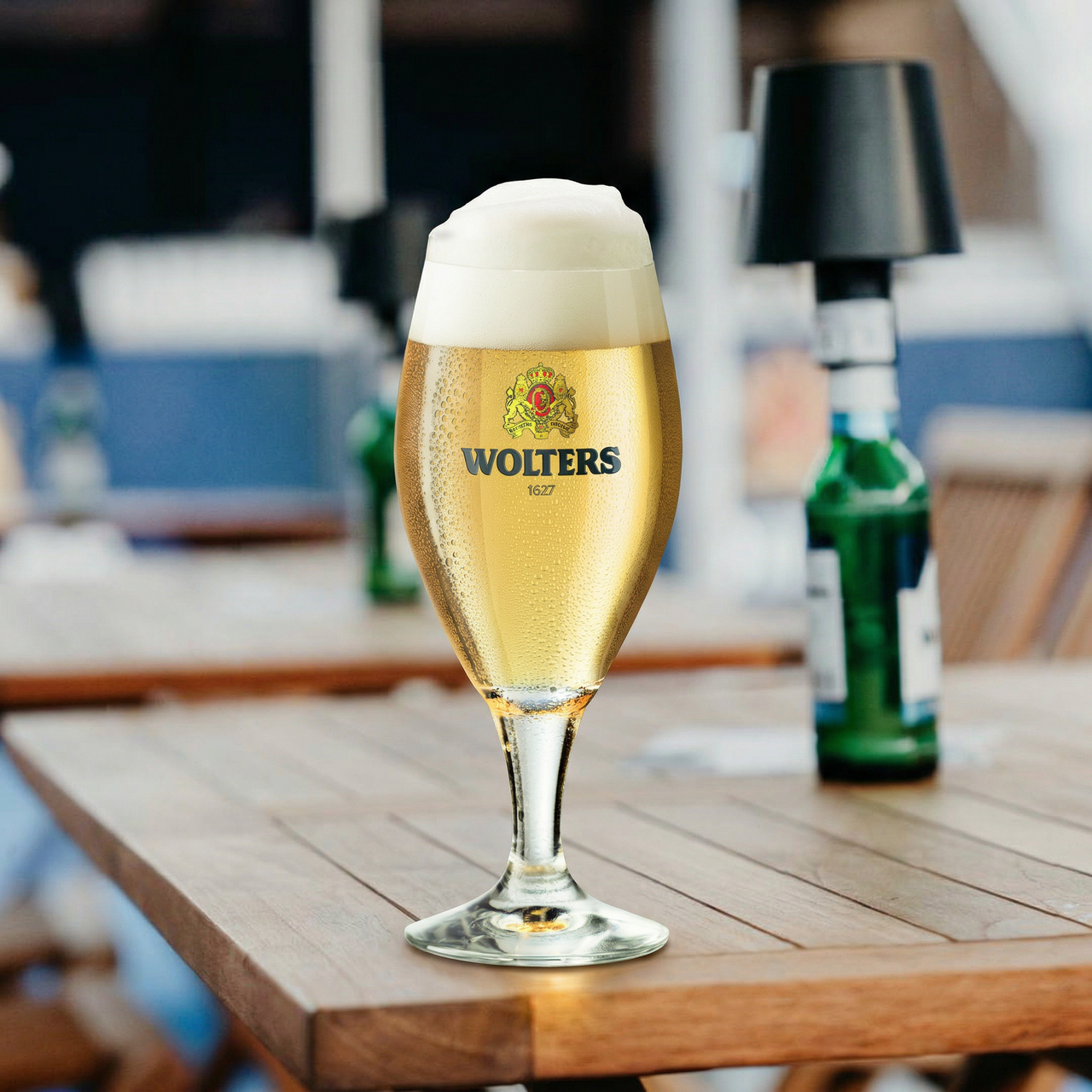 Ein gefülltes Bierglas mit Wolters-Logo steht auf einem Holztisch im Freien. Im Hintergrund ist eine verschwommene Flasche und eine moderne Lampe zu sehen. Das Bier ist hell und hat eine schaumige Krone.