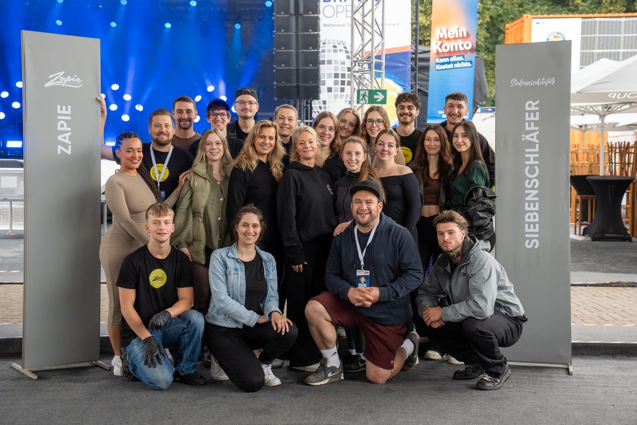 Eine Gruppe von etwa 20 Menschen steht und hockt zwischen zwei großen, vertikalen Schildern, auf denen "Zapie" und "Siebenschläfer" steht. Sie lächeln in die Kamera. Im Hintergrund sind eine Bühne mit blauen Lichtern und Werbebanner sichtbar. Die Gruppe ist in legerer Kleidung, einige tragen Festival-Ausweise um den Hals. Der Ort scheint ein Veranstaltungsbereich im Freien zu sein.