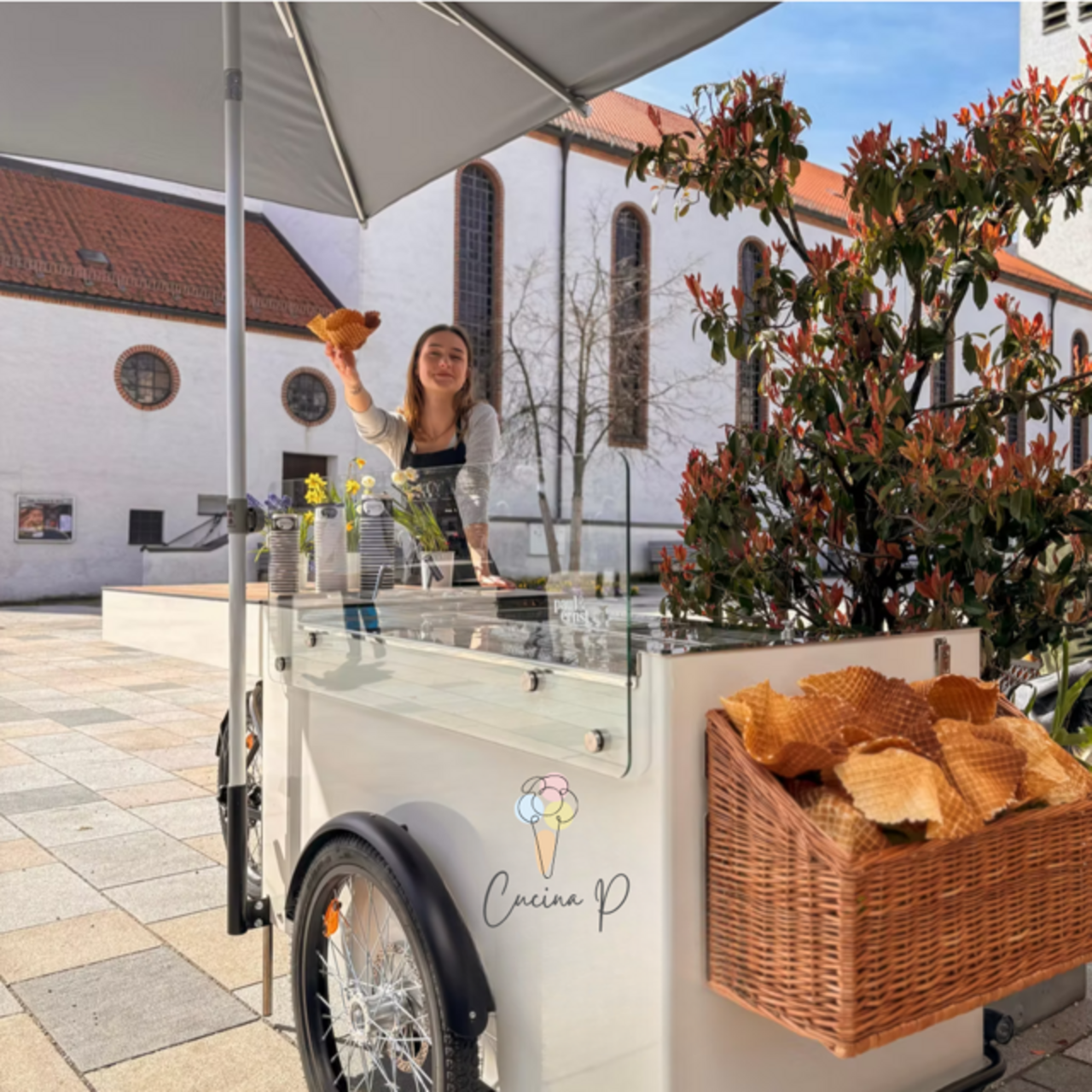 Frau an einem Eis-Bike mit Sonnenschirm, hält eine Waffel, Korb mit Waffeln am Fahrrad, Gebäude im Hintergrund.