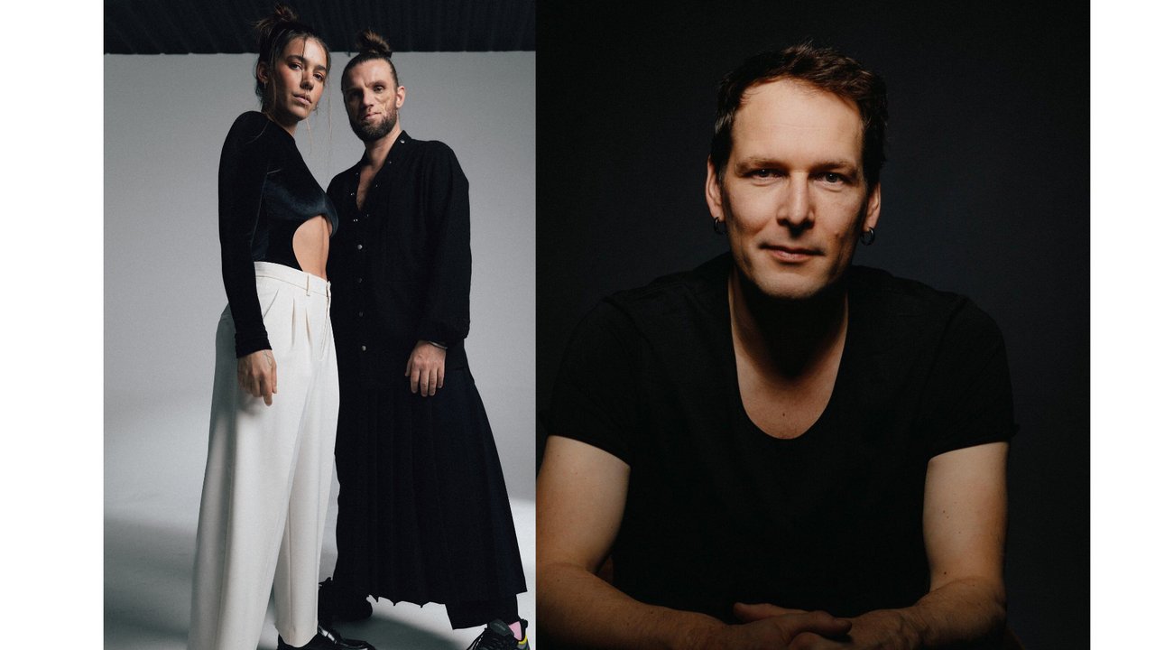 Auf der linken Seite des Bildes befindet sich eine Frau in einem schwarzen Oberteil und weißen Hosen, neben einem Mann, der ebenfalls in schwarz gekleidet ist. Beide stehen in einem Studio. Auf der rechten Seite ist ein Mann in einem schwarzen T-Shirt zu sehen, der mit einem ruhigen Ausdruck auf einem Stuhl sitzt, vor einem dunklen Hintergrund.