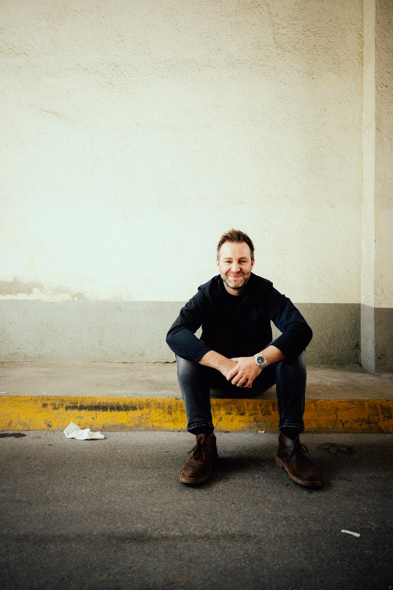 Josh. Ein Mann sitzt auf einem Bordstein vor einer beigen Wand. Er trägt dunkle Kleidung und braune Schuhe, hat kurze Haare und ein freundliches Lächeln. Neben ihm liegt ein Stück Papier auf dem Boden. Die Szene ist in einem leicht schattigen Bereich aufgenommen.
