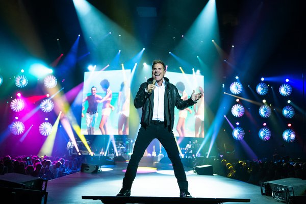 Ein Sänger steht auf einer Bühne bei einem Konzert in einer Arena. Er trägt eine schwarze Lederjacke über einem weißen Hemd und hält ein Mikrofon in der Hand, während er leidenschaftlich singt. Hinter ihm leuchtet ein großer Bildschirm mit einem bunten Bild von tanzenden Menschen am Strand. Die Bühne ist in farbenfrohes Licht getaucht, mit zahlreichen Scheinwerfern, die das Publikum in verschiedenen Farben anstrahlen. Die Stimmung im Publikum wirkt ausgelassen.