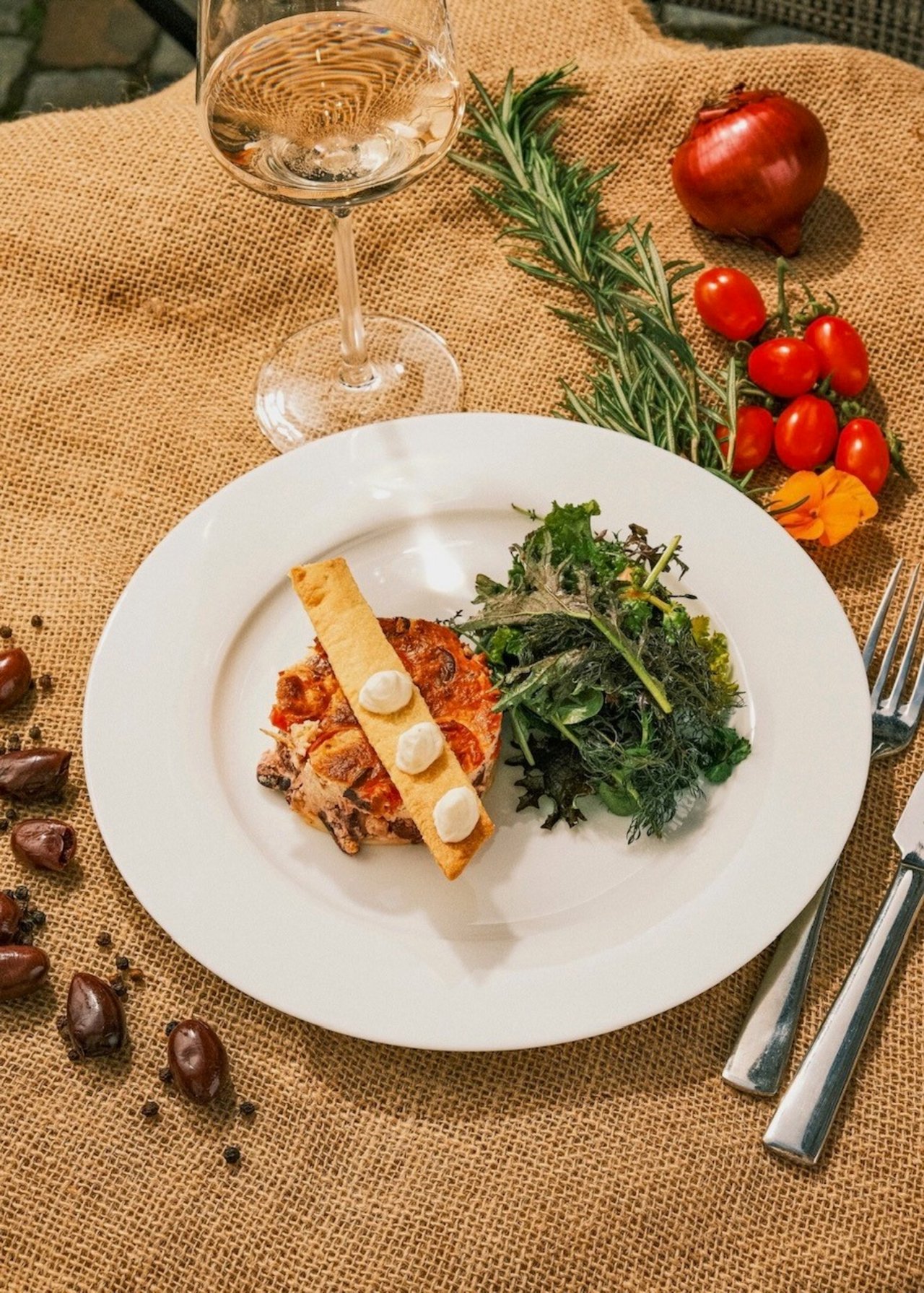 Ein Teller mit einer kleinen Portion Lasagne, daneben ein Stück knuspriges Brot mit drei weißen Tupfen und einem Salat aus frischen Blättern. Im Hintergrund liegt ein Glas Weißwein. Auf dem Jutetuch um den Teller herum sind Rosmarin, Tomaten, eine rote Zwiebel, Datteln und ein paar Pfefferkörner arrangiert.