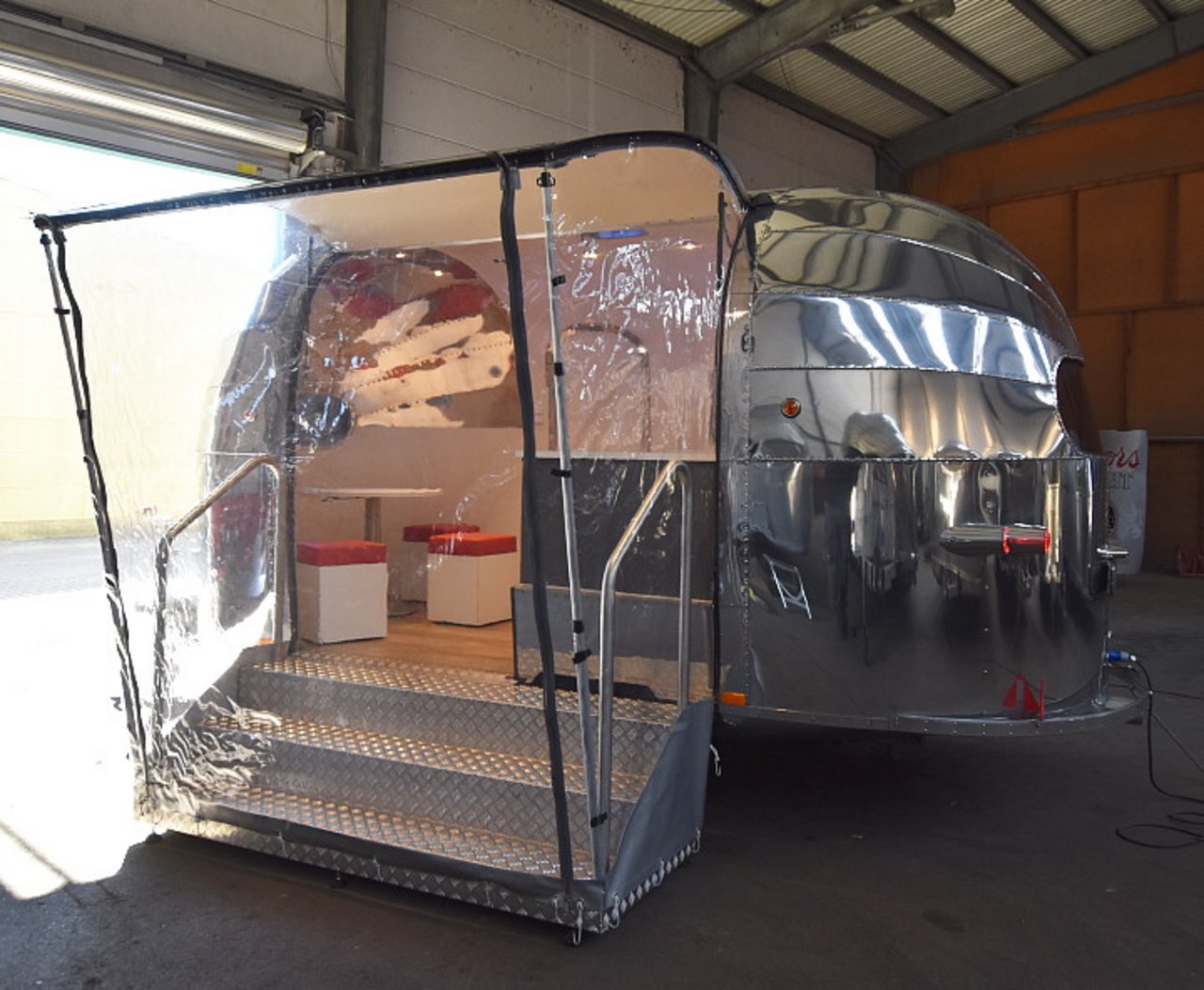 Ein silber glänzender Wohnwagen mit einem überdachten Eingangsbereich aus transparentem Plastik. Der Eingangsbereich hat Treppen mit Metallgeländern und führt zu einer kleinen Sitzecke im Inneren des Wohnwagens. Die Innenausstattung scheint modern und hell, mit roten und weißen Akzenten. Der Wohnwagen steht in einer großen Halle.