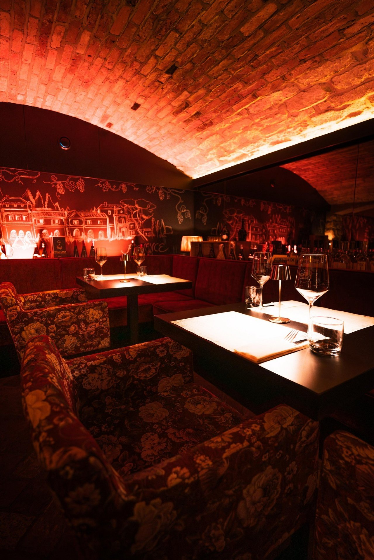 Ein gemütliches Restaurant mit stimmungsvoller Beleuchtung. Die Backsteinwände sind rot beleuchtet und sorgen für eine warme Atmosphäre. Zwei Tische sind mittig ausgerichtet, gedeckt mit Gläsern und Besteck. Die Stühle sind mit einem floralen Muster bezogen. Ein Wandgemälde zeigt eine stilisierte Landschaft mit Gebäuden.