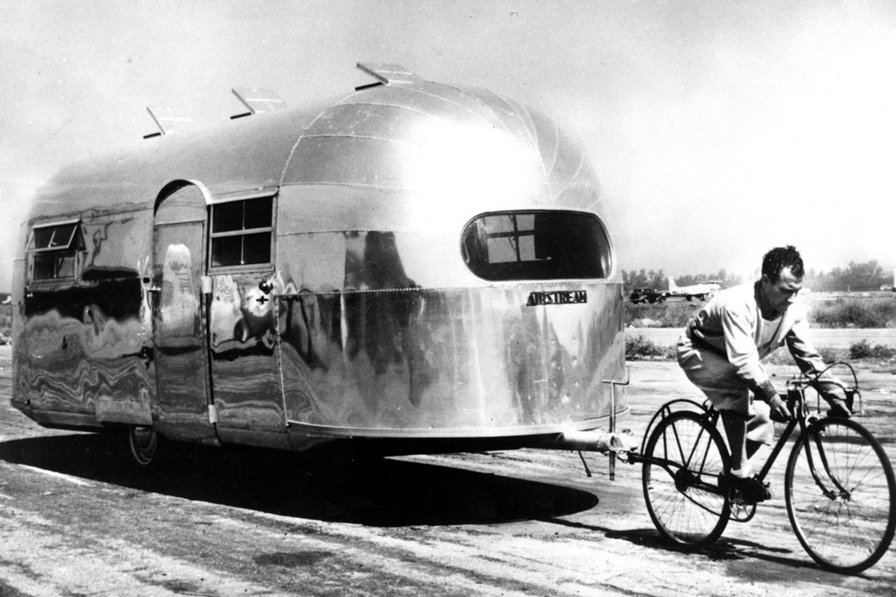 Ein Mann fährt ein Fahrrad und zieht dabei einen großen, stromlinienförmigen Wohnwagen aus glänzendem Metall hinter sich her. Der Wohnwagen wirkt futuristisch und reflektiert das Sonnenlicht. Die Szene scheint aus einer vergangenen Ära zu stammen, erkennbar an der alten Fahrrad- und Wohnwagentechnologie. Der Hintergrund ist weitgehend offen und zeigt eine unbefestigte Straße und einen wolkenlosen Himmel.