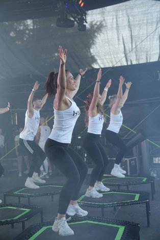 Eine Gruppe von Personen in weißen Sportoberteilen und schwarzen Leggings führt eine Fitnessroutine auf Trampolinen durch. Alle befinden sich in einem synchronen Sprung mit erhobenen Armen. Im Hintergrund sind Bühnenelemente und Lichttechnik sichtbar. Die Szenerie wirkt wie eine Aufführung oder ein Fitness-Event.