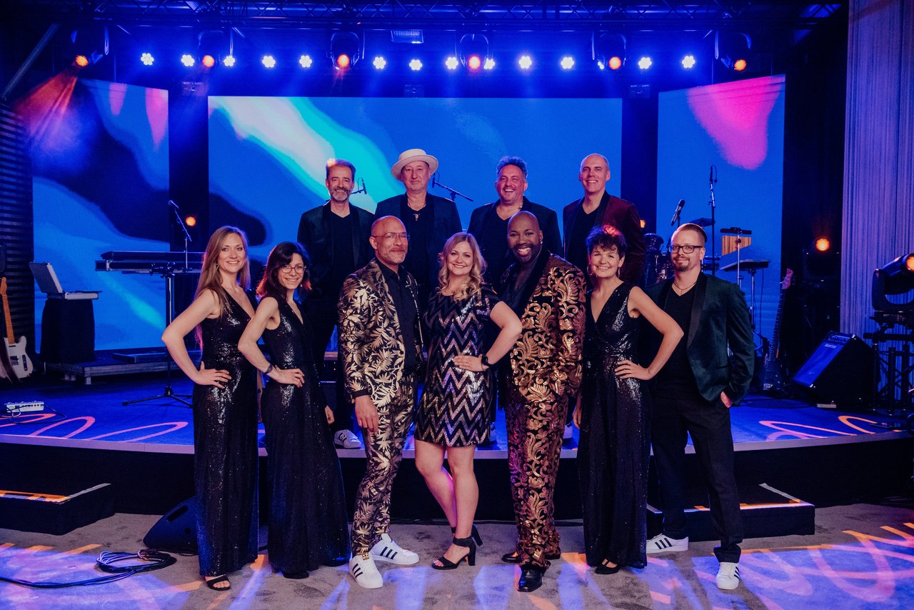 Watch your Steps Eine Gruppe von elf Personen steht lächelnd auf einer Bühne vor einer farbenfrohen Hintergrundbeleuchtung. Sie tragen festliche Kleidung, darunter glitzernde Kleider und gemusterte Anzüge. Im Hintergrund sind Instrumente und Mikrofone zu sehen. Die Atmosphäre wirkt fröhlich und festlich.