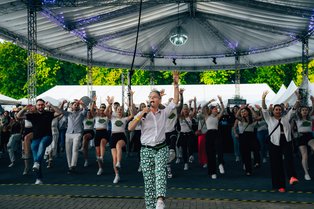 Eine Gruppe von Menschen tanzt unter einem großen, überdachten Pavillon. Im Vordergrund steht eine Person mit einem Mikrofon in der Hand, die bunte, gemusterte Hosen trägt. Die Menge im Hintergrund folgt enthusiastisch den Tanzbewegungen mit erhobenen Armen. Die Veranstaltung findet draußen bei Tageslicht statt, und es gibt mehrere weiße Zelte im Hintergrund. Die Atmosphäre wirkt fröhlich und mitreißend.