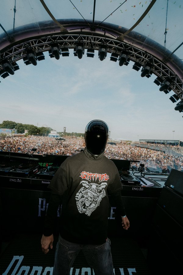 Eine Person mit einem schwarzen, gesichtsverdeckenden Helm steht auf einer Bühne bei einem Musikfestival. Sie trägt ein dunkles T-Shirt mit einem auffälligen Grafikdruck. Hinter der Bühne erstreckt sich eine große Menschenmenge unter freiem Himmel. Über der Bühne befindet sich eine beeindruckende Lichtkonstruktion. Der blaue Himmel ist im Hintergrund sichtbar.