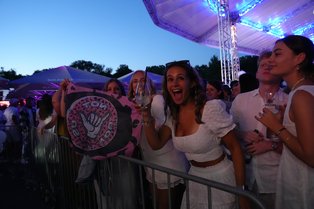 Eine Gruppe junger Menschen auf einem Event steht hinter einer Absperrung. Eine Frau in der Mitte trägt ein weißes Kleid und hält ein Glas, schaut freudestrahlend in die Kamera. Neben ihr sind weitere Personen, die ebenfalls Getränke in den Händen halten. Die Stimmung ist ausgelassen. Über ihnen hängt eine beleuchtete Überdachung, und im Hintergrund sind Bäume und Schirme zu sehen. Ein auffälliges Schild mit einer Handgeste ist im Vordergrund.