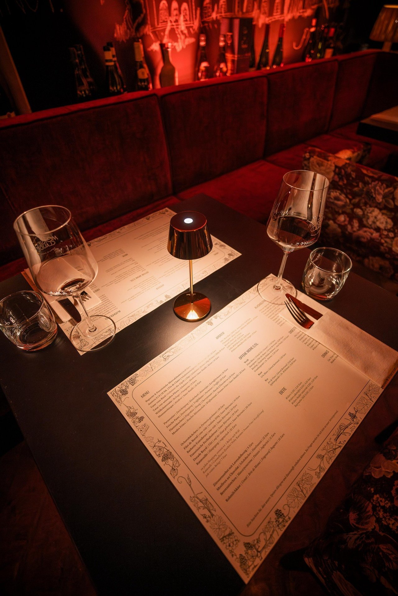 Das Bild zeigt einen stilvoll gedeckten Tisch in einem Restaurant mit gedämpfter Beleuchtung. Auf dem Tisch liegen zwei Menükarten, flankiert von Weingläsern und Wassergläsern. Eine kleine Tischlampe mit warmem, goldenen Licht steht zwischen den Karten. Der Hintergrund besteht aus einer weinroten Polsterbank, und die Wand ist mit Weinflaschen dekoriert, die von einem sanften roten Licht angestrahlt werden.