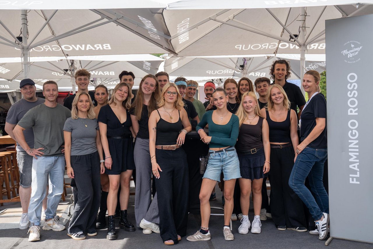 Auf dem Bild ist eine Gruppe von Menschen zu sehen, die unter weißen Sonnenschirmen steht. Sie lächeln und posieren für die Kamera. Die Personen tragen überwiegend legere Kleidung in gedeckten Farben. Im Hintergrund sind Tische und Stühle zu erkennen. Rechts im Bild ist ein Schild mit der Aufschrift "Flamingo Rosso" sichtbar.