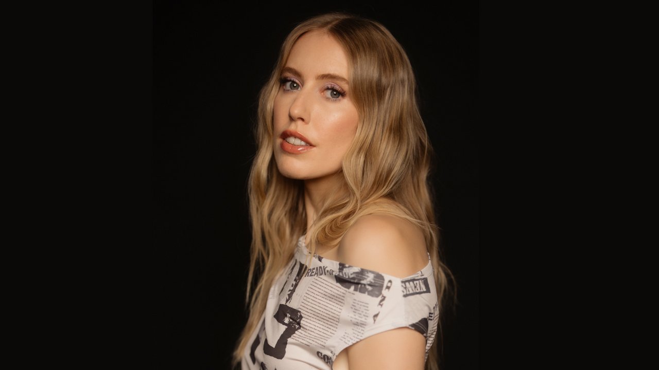 Das Bild zeigt eine Frau mit langen, welligen, blonden Haaren, die zur Seite schaut. Sie trägt ein Oberteil mit einem Muster aus Zeitungsdruck. Der Hintergrund ist schwarz, was die Person hervorhebt.