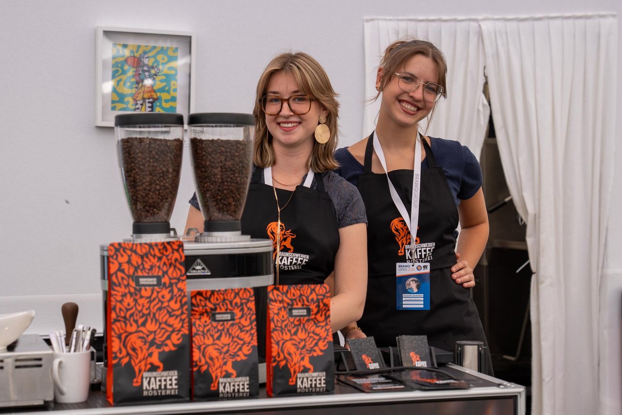 Zwei Personen stehen lächelnd an einem Stand. Sie tragen schwarze Schürzen mit dem Logo einer Kaffeerösterei. Vor ihnen sind mehrere Packungen Kaffee und zwei große Kaffeemühlen zu sehen. Im Hintergrund hängt ein buntes Bild an der Wand und ein weißer Vorhang ist zur Seite geschoben.
