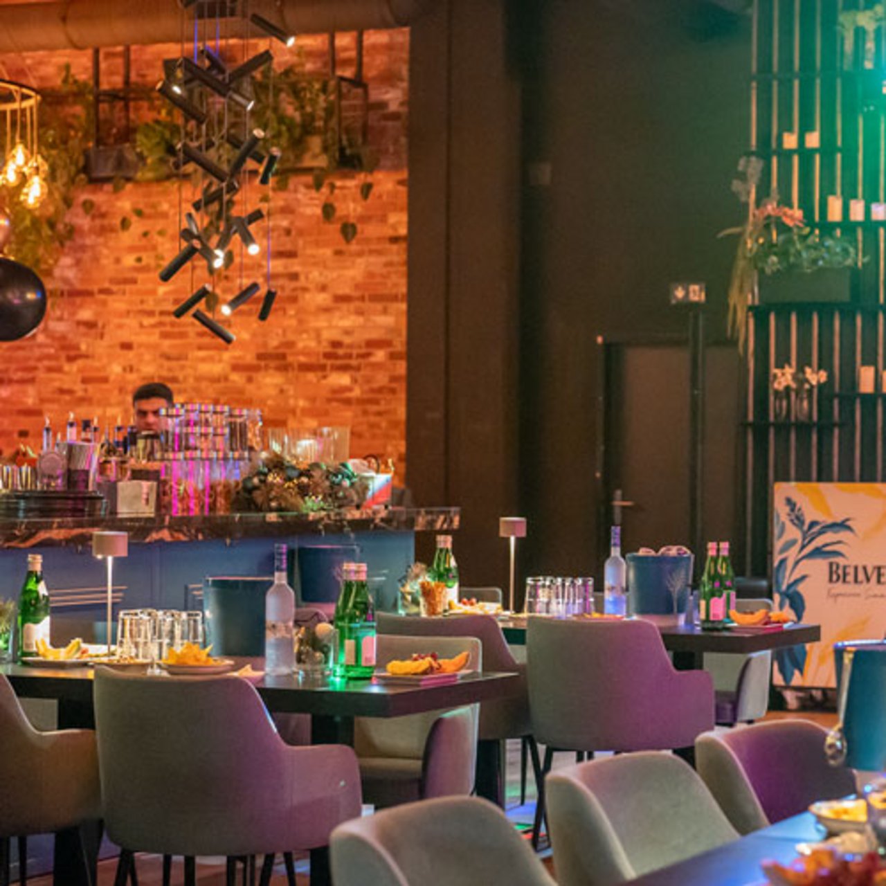 Das Bild zeigt ein stilvoll eingerichtetes Restaurant oder Café mit modernen Möbeln und einem gemütlichen Ambiente. Die Tische sind mit Getränken und Gläsern versehen, während im Hintergrund eine Bar mit dekorativen Elementen wie Pflanzen und Beleuchtung zu sehen ist. An der Wand befindet sich eine Backsteinoptik, die dem Raum einen industriellen Touch verleiht. Die Beleuchtung ist warm und einladend.