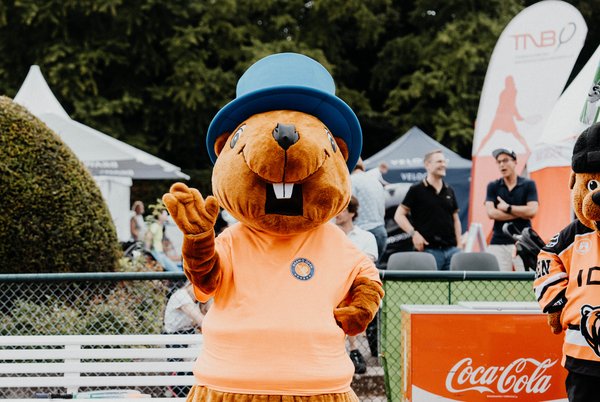 Ein großes Maskottchen in Form eines Bibers trägt ein orangefarbenes T-Shirt und einen blauen Hut. Es steht auf einem Tennisplatz-Event und winkt in die Kamera. Im Hintergrund sind Zuschauer und diverse Stände mit Werbematerialien zu sehen. Auf einem der Stände ist das Coca-Cola-Logo prominent platziert.